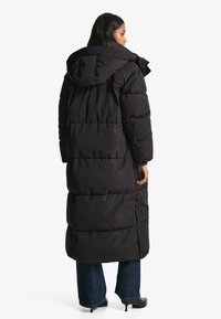 Geacă matlasată lungă neagră cu glugă, având un design matlasat, secțiuni umflate și un guler ajustabil. Poartă peste blugi de denim închis și tocuri negre.