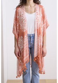 Perzikkleurig poncho-stijl kledingstuk met witte, ingewikkelde patronen, franjes aan de zoom, lichtgewicht stof, open voorkant, gedragen over een witte top en spijkerbroek.