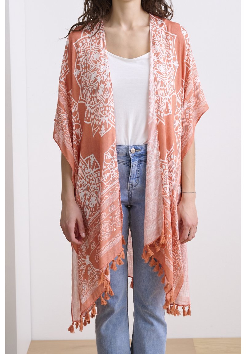 Perzikkleurig poncho-stijl kledingstuk met witte, ingewikkelde patronen, franjes aan de zoom, lichtgewicht stof, open voorkant, gedragen over een witte top en spijkerbroek.