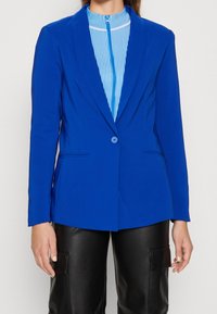 Klart blå skräddarsydd kavaj med lång ärm, enkel knappknäppning och två framfickor. Bärs över en blå och vit randig topp.