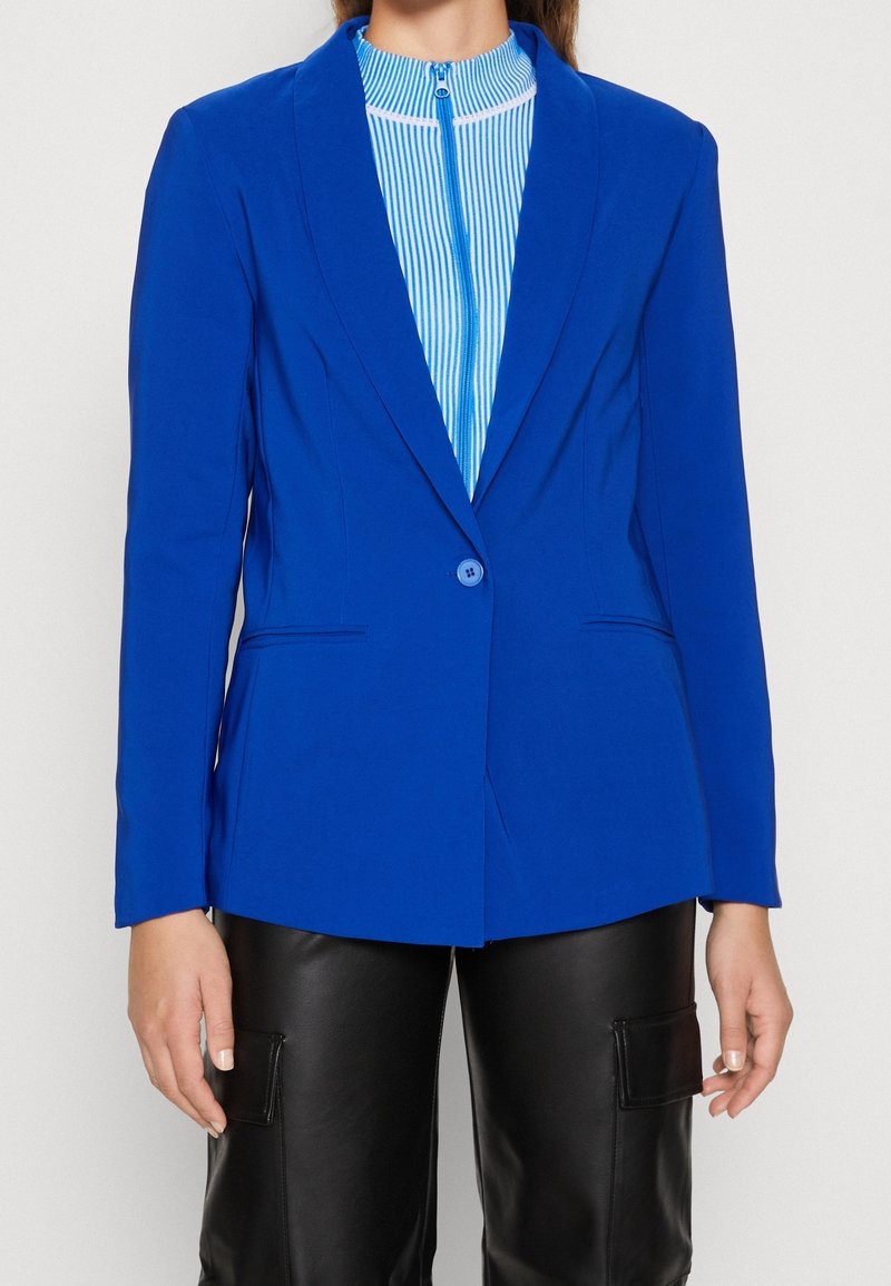 Klart blå skräddarsydd kavaj med lång ärm, enkel knappknäppning och två framfickor. Bärs över en blå och vit randig topp.