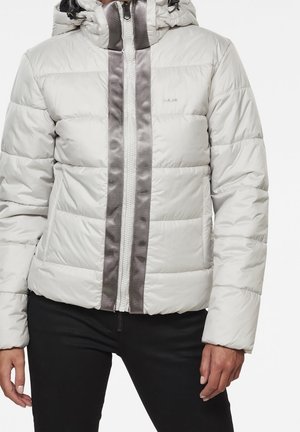 Person trägt eine hellgraue Steppjacke mit Frontreißverschluss und schwarze Hose, Hände entspannt an den Seiten.