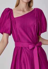 Robe fuchsia avec des manches bouffantes, un décolleté asymétrique et une ceinture nouée à la taille. Tissu lisse et texturé avec un léger éclat.