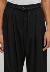 Schwarze Plisséhose mit einem taillierten Bund, Stoffgürtel mit D-Ring und schlanker Passform. Glatte Textur ohne sichtbare Muster.