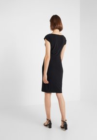 Žena s krátkými hnědými vlasy, oblečená v přiléhavých černých šatech ke kolenům a černých sandálech na podpatku, stojící u bílé rohové zdi.