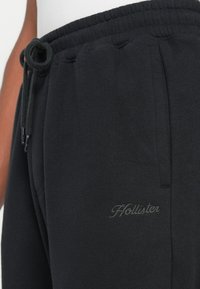 Krupan plan crnih trenirki s podesivim strukom, bočnom džepom i diskretnom izvezenom Hollister logotipom na bedru.
