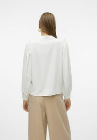 Blusa bianca con dettagli arricciati sulle spalle, maniche lunghe e polsini con bottoni, abbinata a pantaloni beige a gamba larga. Texture liscia e tonalità neutre.