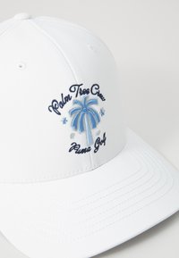 Hvit baseballcap med blå broderi av et palmetre og teksten "Palm Tree Crew" og "Puma Golf." Glatt stoff med sydd detaljer.