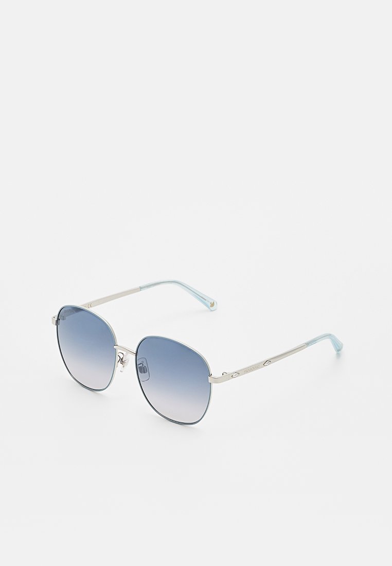Lunettes de soleil avec cadre argenté, verres dégradés bleus et embouts de branches bleu clair, reposant sur une surface blanche.