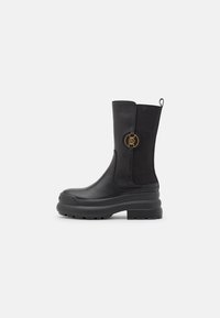Black ankle boots made of smooth leather with a chunky rubber sole, featuring a gold circular accent and elastic side panels for comfort.