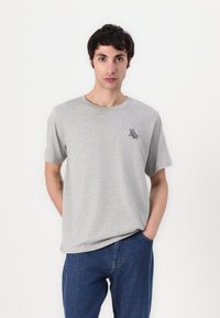 Tricou gri cu mânecă scurtă, dintr-un material ușor; are o grafică mică bleu marin pe partea stângă a pieptului și un guler clasic rotund.