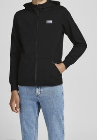 Man som bär en svart zip-up hoodie med logotyp patch på bröstet och ljusblå jeans, ena handen i fickan, mot en neutral bakgrund.