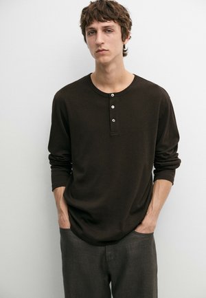 Ung mand med lyst brunt kruset hår iført en mørkebrun langærmet henley-shirt og grå bukser, stående med hænderne i lommerne.