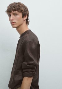 Dark brown, long-sleeve shirt with a round neck and rolled cuffs. Fabric appears soft with subtle texture. Model poses in profile against a light background.