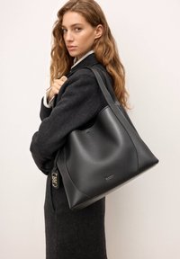 Sort læderhåndtaske med et glat, struktureret design og afrundede kanter, der har en metallisk hardwaredetalje og brandlogo.