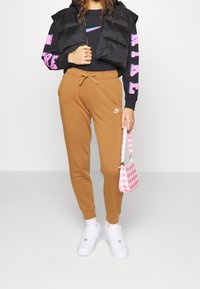 Svart puffväst över en grafisk svart sweatshirt, tan joggers med snörning, vita sneakers och en rosa rutiga handväska.