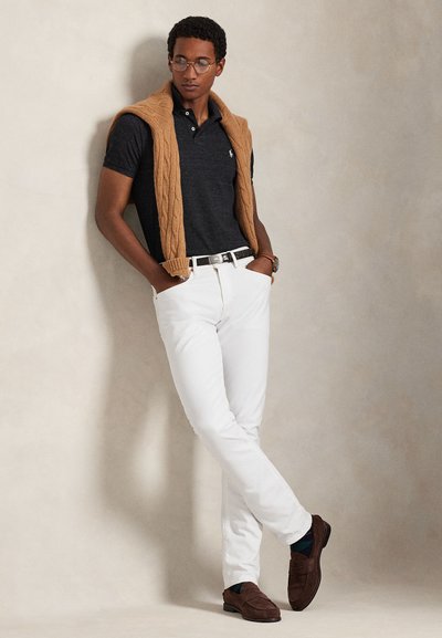Man wearing glasses, a black polo shirt, white pants, brown shoes, and a brown sweater draped over shoulders, standing with hands in pockets.