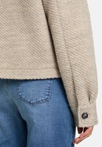 Pull beige texturé avec des côtes diagonales, présentant un détail de poignet boutonné. Assorti à un jean en denim bleu, mettant en avant le design de la poche arrière.