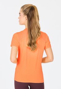 Kortärmad, åtsittande träningsskjorta i klar orange, med en mjuk textur och ventilerad design vid ryggen, matchad med mörka leggings.