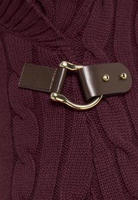 Gros plan sur une boucle en cuir marron avec une ferrure dorée fixée à un tissu en maille torsadée de couleur bordeaux foncé.