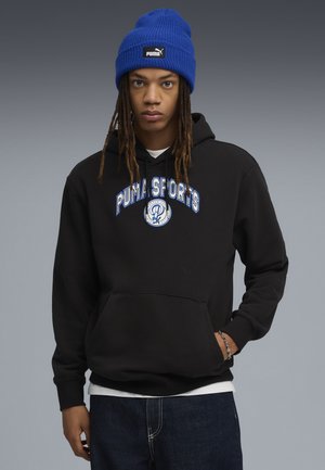 Svart hoodie med en framficka och blå "PUMA SPORTS" logotyp. Bärs med en blå stickad mössa. Mörka jeans fulländar outfiten.
