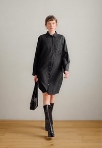 Μαύρο τζιν φόρεμα με μακριά μανίκια, κουμπιά μπροστά και τσέπη στο πλάι. Φοριέται με μαύρες μπότες μέχρι το γόνατο και μαύρη τσάντα ώμου.