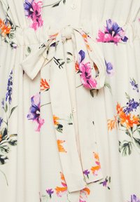 Vestido floral en crema con estampados en púrpura, naranja y rosa. Presenta una tela texturizada, un cierre de botones en la parte delantera y un cinturón anudado a la cintura.