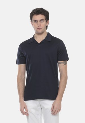 Giovane uomo con capelli corti e scuri, che indossa una camicia nera a maniche corte con colletto e pantaloni bianchi, in piedi davanti a uno sfondo bianco uniforme.