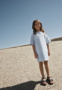 Světlomodré bavlněné šaty s vyšívaným lemováním ve vlnkovém vzoru a rukávy připomínající krajku, doplněné černými sandály s přezkami.