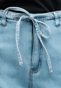 Φωτεινός γαλάζιος τζιν παντελόνι με κορδόνι από ύφασμα δεμένο σε φιόγκο στη μέση, εμφανείς θηλιές για ζώνη και αντίθετη πορτοκαλί ραφή.