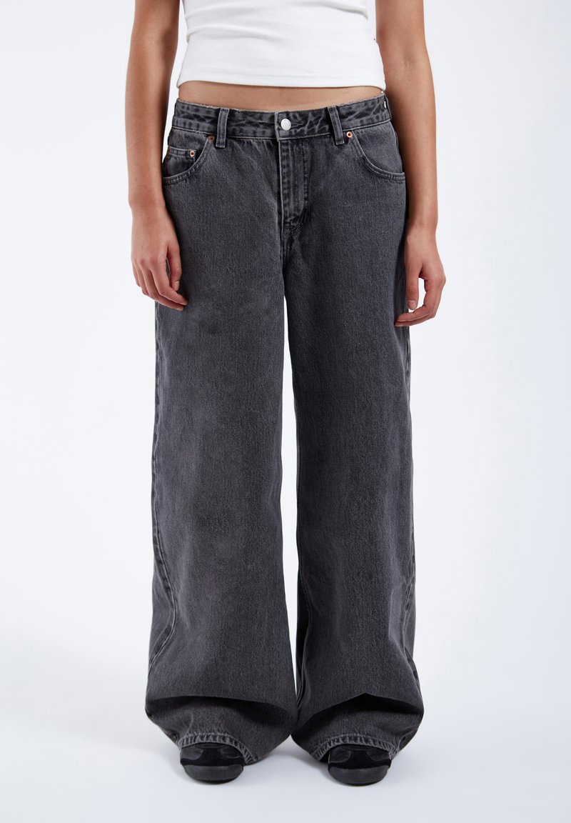 Persoon draagt losse zwarte denim jeans en een wit cropped top, staand tegen een effen lichte achtergrond.