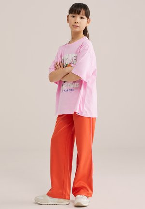 Jauna mergina su tamsiais plaukais surištais į kuoduką, dėvinti laisvą rožinę megztinį su grafiniu dizainu, ryškias oranžines plačias kelnes ir baltus sportinius batelius, stovinti sukryžiavusi rankas.