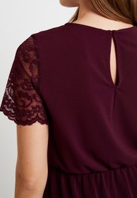 Femme portant une robe violet foncé avec des manches courtes en dentelle et une petite ouverture en forme de trou de serrure au dos du col.