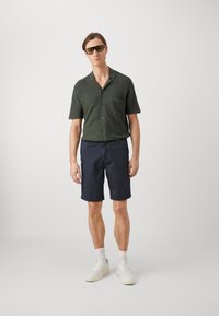 Camicetta verde a maniche corte con bottoni e tasca, abbinata a pantaloni corti blu. Modello che indossa scarpe da ginnastica bianche e occhiali da sole con lenti verdi.