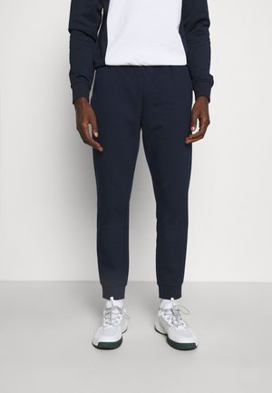 Mann trägt marineblaue Jogginghosen, ein weiß-blaues Sweatshirt und weiße Turnschuhe vor einem schlichten hellen Hintergrund.