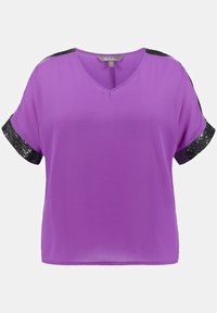 Lila blus med V-ringning och korta ärmar. Ärmarna har svarta paljettaccenter. Tillverkad av ett lätt, mjukt tyg.