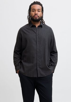 Homme avec barbe et dreadlocks, debout face à nous, portant une chemise boutonnée gris foncé et un pantalon noir, les mains dans les poches.