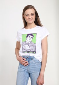 Bílá tričko s grafikou svalnaté postavy a textem "THE HULK" na zeleném pozadí. Krátké rukávy, kulatý výstřih.