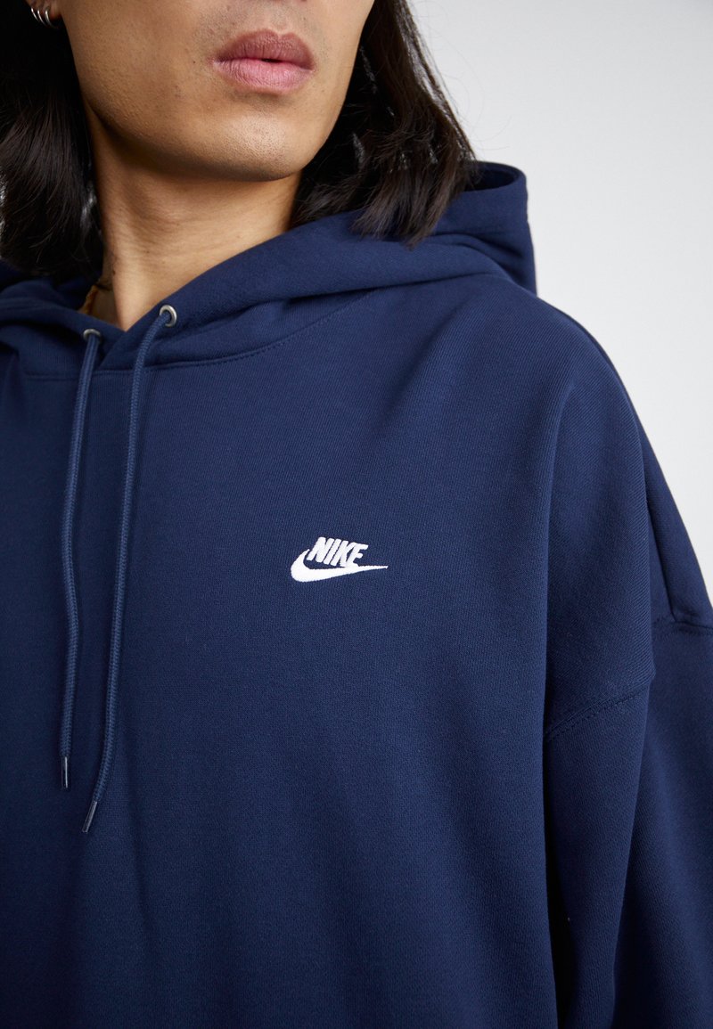 Granatowy hoodie wykonany z miękkiego materiału, z kieszenią typu kangur i białym logo Nike na piersi. Bluza z kapturem z regulowanymi sznurkami.