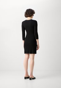Robe noire côtelée à manches trois-quarts, au design ajusté, atteignant le mi-cuisse. Complétée par des mules à talons blocs noirs. Fini lisse et texturé.