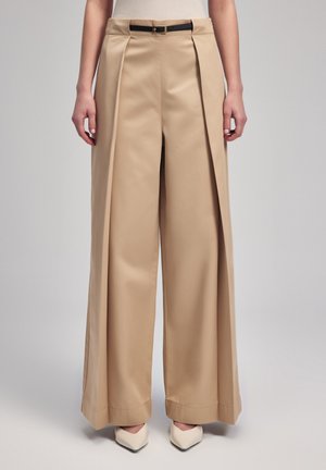 Beige weite Hose mit Falten an der Vorderseite, einem schwarzen Gürtel und glatter Stoffoberfläche, kombiniert mit hellen spitzen Absätzen.