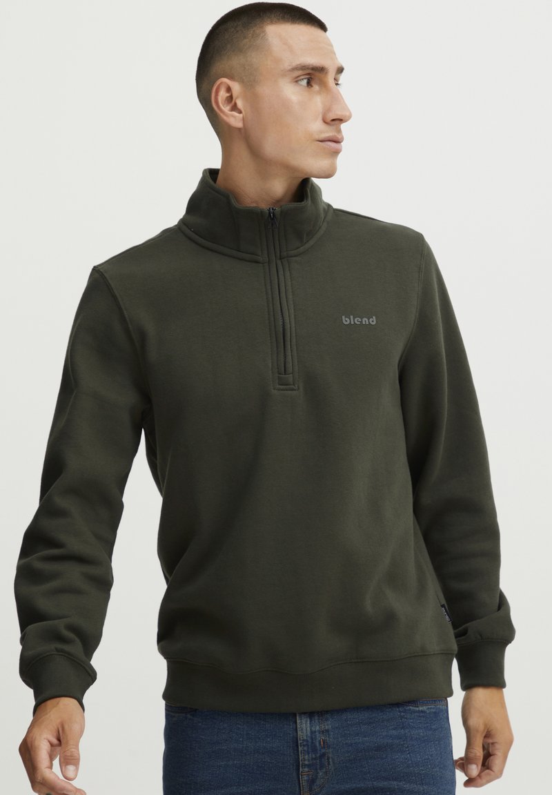 Sudadera de color verde oliva con cremallera de cuarto, textura suave, puños acanalados y logo en el pecho; ajuste casual.