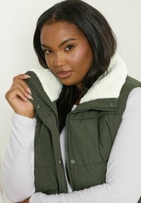 Zielona kamizelka puchowa z białym polarowym kołnierzem, zapinana na zamek błyskawiczny i zatrzaskami, zestawiona z białą koszulą z długim rękawem. Gładka faktura materiału.