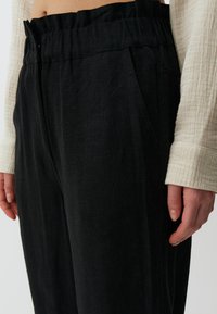 Schwarze, locker sitzende Hose mit elastischem Bund, seitlichen Taschen und einem weichen, strukturierten Stoff, kombiniert mit einem hellen Hemd.