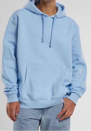 Man draagt een lichtblauwe hoodie met voorzak en trekkoorden, gecombineerd met lichtblauwe spijkerbroek, tegen een effen achtergrond.