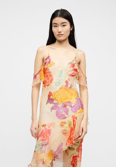 Νεαρή γυναίκα με μακριά σκούρα μαλλιά που φοράει μια διάφανη, φορεσιά με λουλουδάτο μοτίβο και μανίκια τύπου «κρύους ώμους» μπροστά σε απλό φόντο.