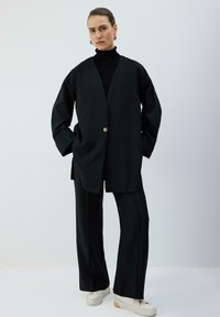 Melns pārmērots žakete ar v-veida izgriezumu, vienu zelta pogu, kombinēts ar melnām platām biksēm un baltām sporta kurpēm. Gluda auduma tekstūra.
