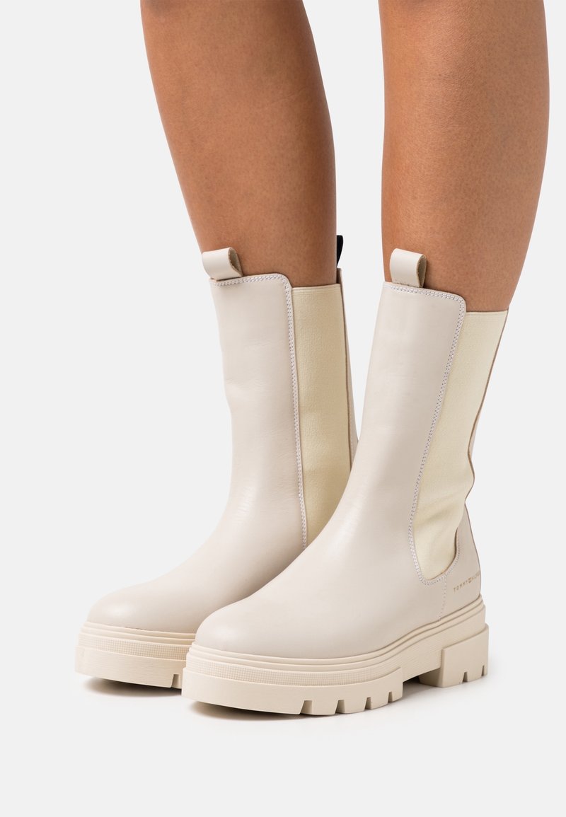 Botas de tobillo de cuero beige con paneles laterales elásticos y una suela de goma gruesa y texturizada. Pestañas en la parte superior para facilitar su uso.
