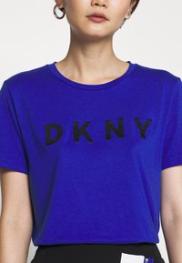 Krótki rękaw, t-shirt z okrągłym dekoltem w intensywnym niebieskim kolorze, z odważnym czarnym napisem "DKNY" na piersi, ozdobionym teksturowanym wykończeniem.