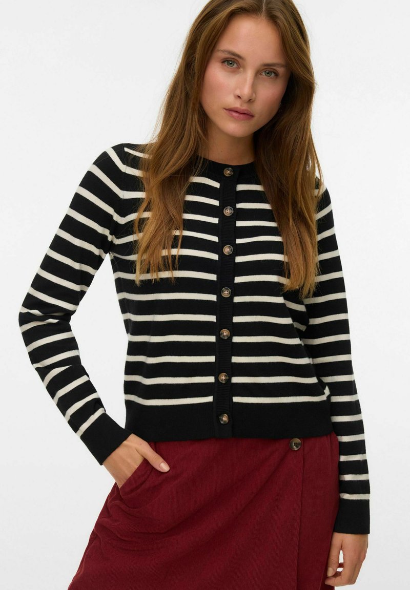 Kvinna iklädd en svart och vit horisontellt randig knäppt cardigan med långa ärmar och en röd kjol med knappdetalj.
